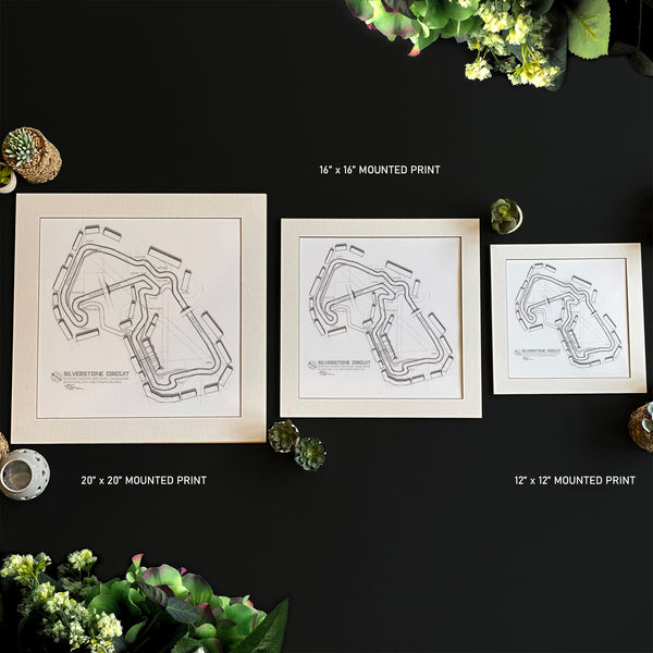 Three framed race track art prints in different sizes (12x12, 16x16, and 20x20 mounted prints) arranged on a dark background with decorative plants and coffee cups.