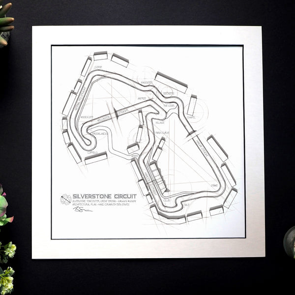 A framed black and white technical circuit map print of Silverstone Circuit, displayed on a dark textured background with small plant leaves at the edges.